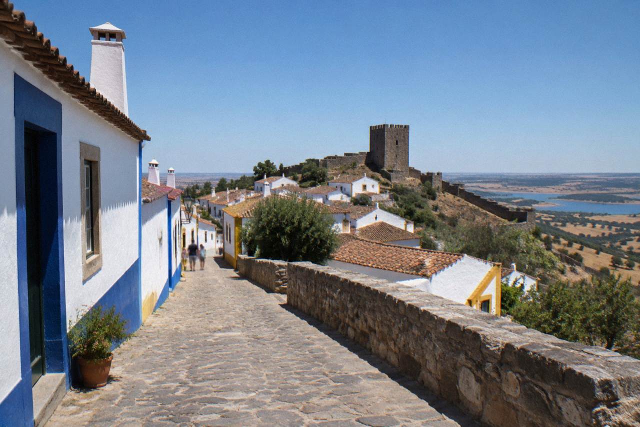 Wide golden plain of the Alentejo with cork oaks and a whitewashed hilltop village under a deep blue sky
