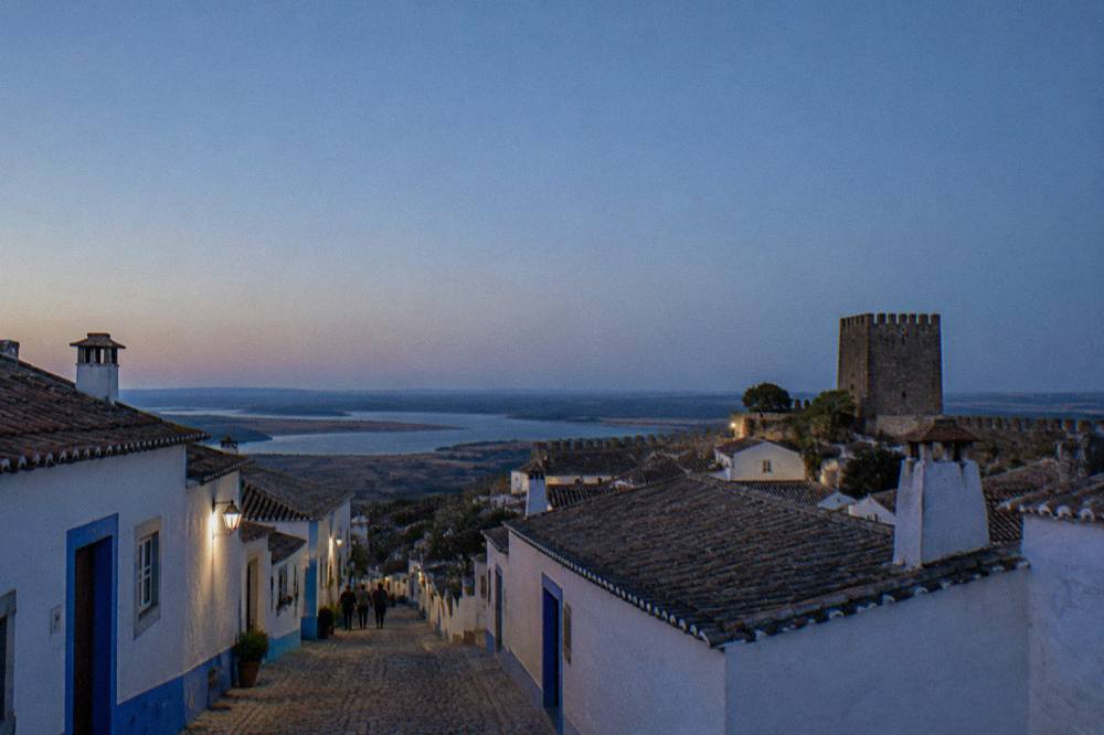 A narrow village street of whitewashed houses with blue trim leading toward a stone castle keep