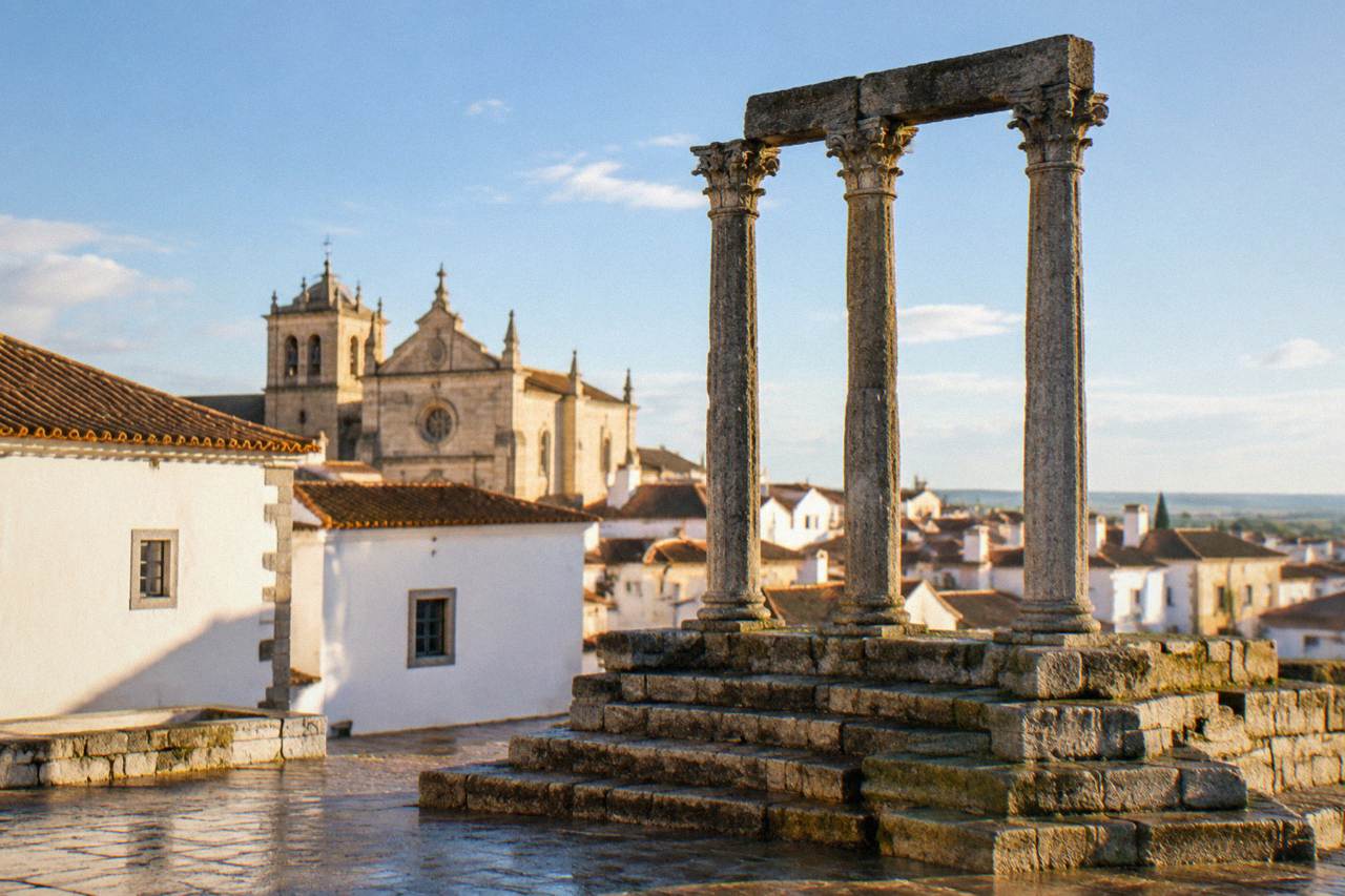 Golden late-afternoon light on the whitewashed houses and tiled roofs of Évora's old town, with the Templo Romano columns rising above the rooftops.