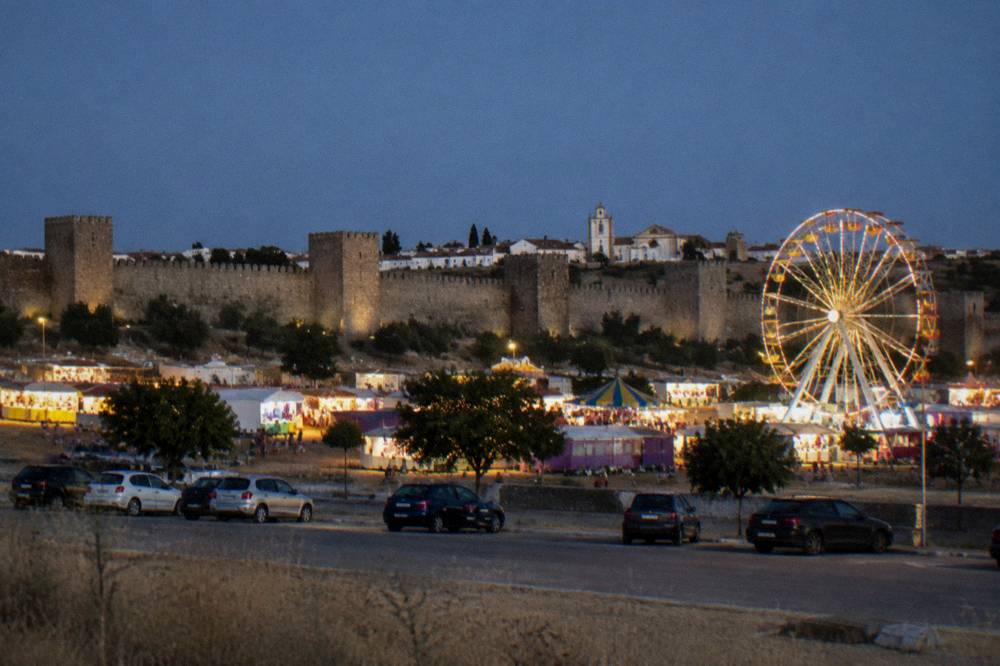 A fairground at dusk with rows of lit stalls and a ferris wheel against a deep blue sky