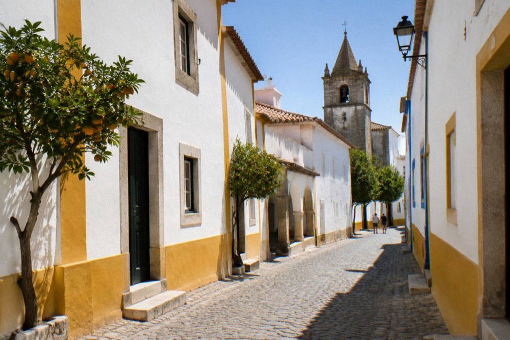 Sunlit stone street in Évora's old town with white-washed walls and a church tower in the background