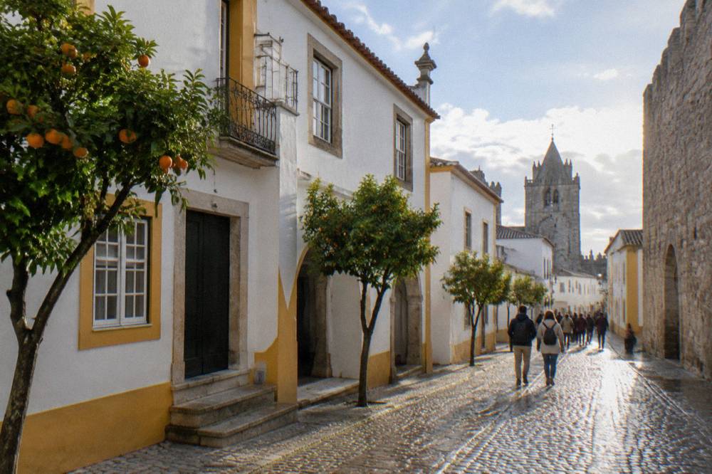 Visitors walking past whitewashed houses with yellow trim near a palace wall in Évora