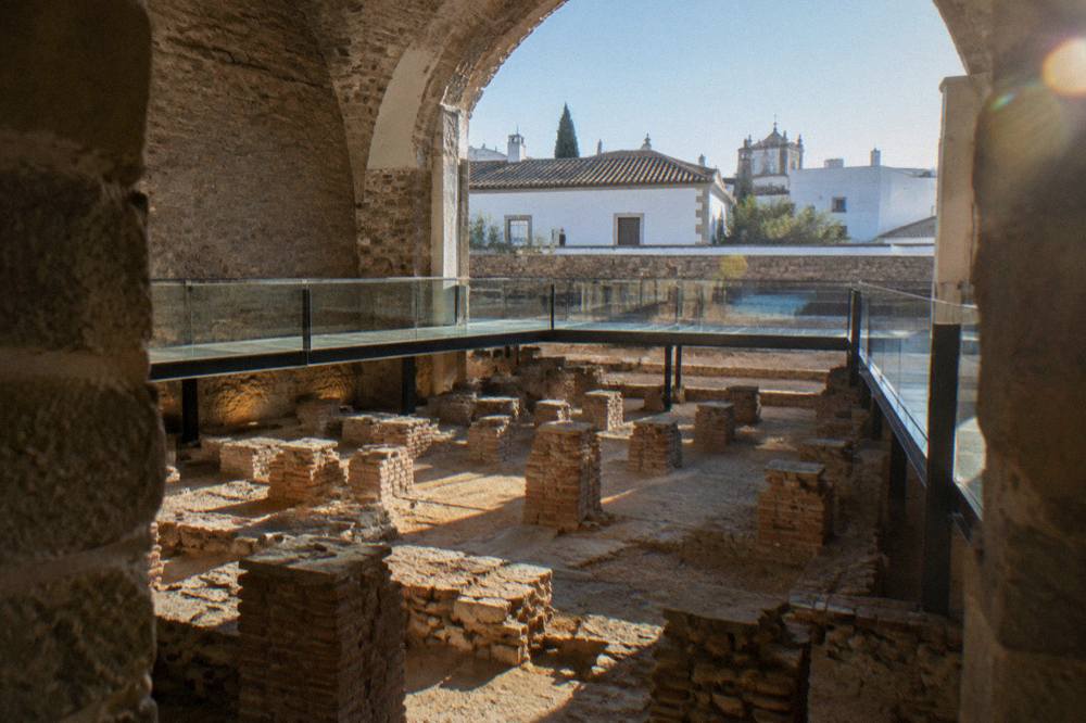 Sunlit ancient stone blocks of a Roman bathhouse foundation with whitewashed Alentejo buildings behind