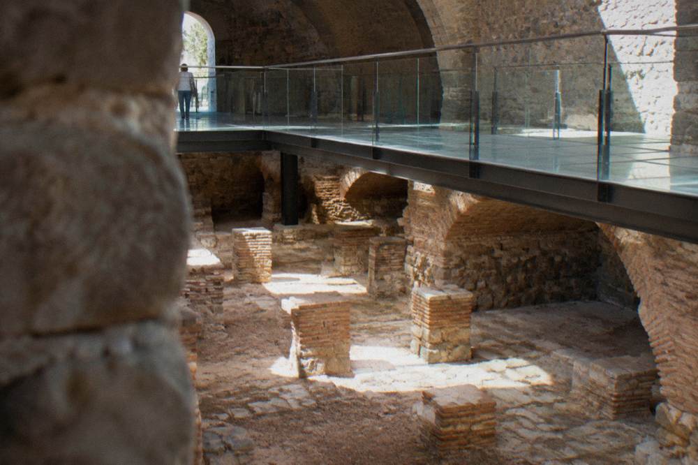 Low ancient arch and stone footing of a Roman bathhouse with a modern pedestrian street passing beside it