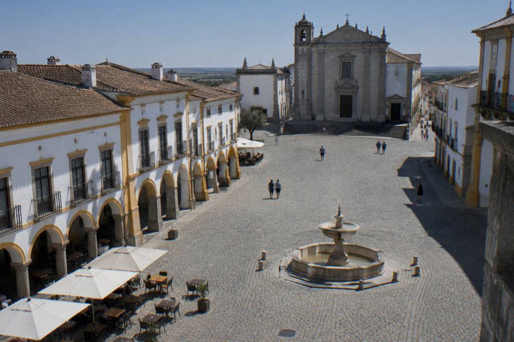 A stone fountain and arched arcades along one side of a historic square in the Alentejo