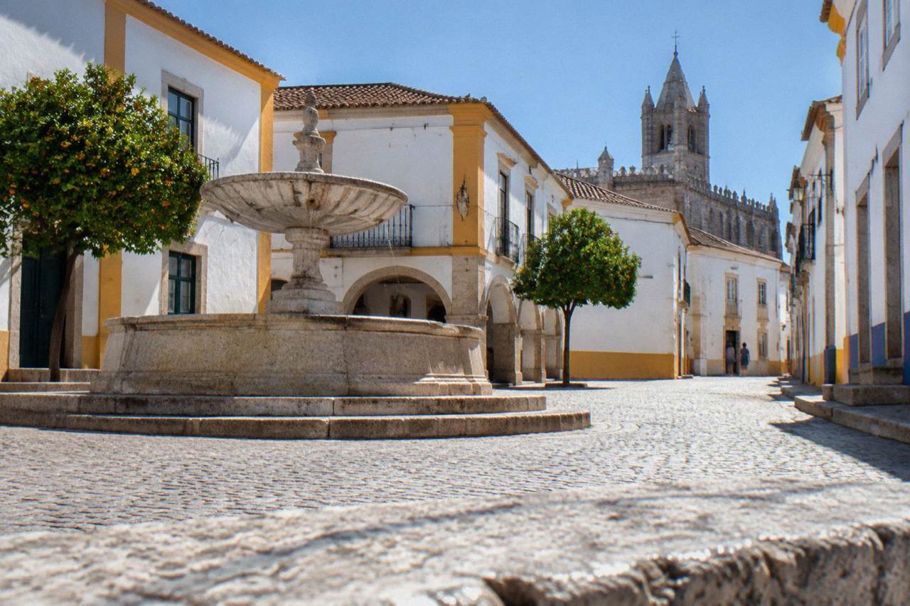 A quiet Renaissance square in Évora with a stone fountain, whitewashed houses and the tower of the university church beyond