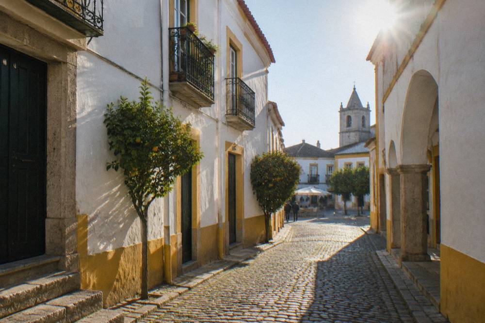 A narrow cobbled lane in Évora's old town with whitewashed walls, yellow trim and iron balconies leading toward a small square