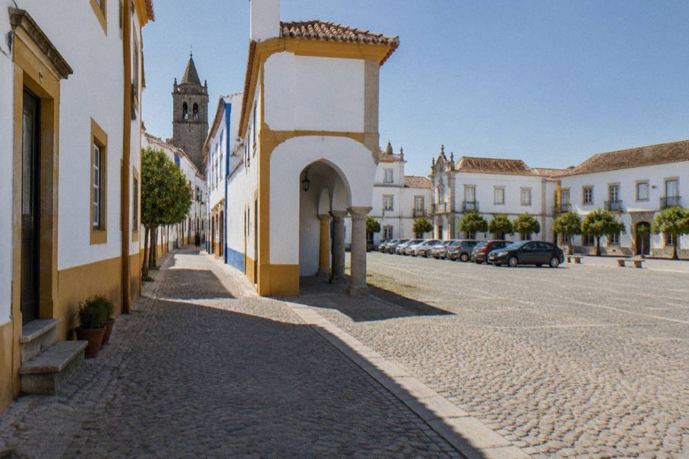 A broad empty esplanade in Évora with parked cars and old buildings along one side under a wide sky