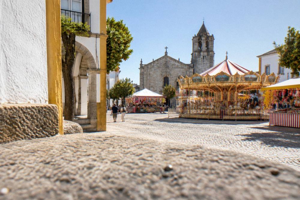 A fairground with stalls and lights set up on a large open square beside a stone chapel in Évora