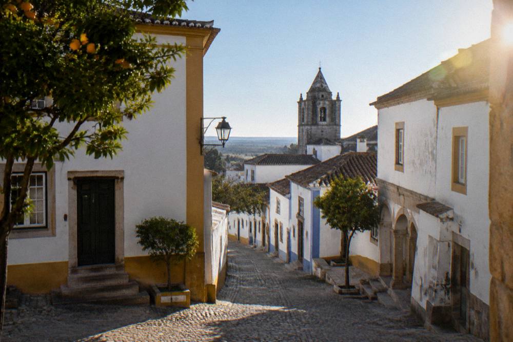 A quiet street corner in Évora with a small stone chapel tower above the rooftops in late afternoon sun
