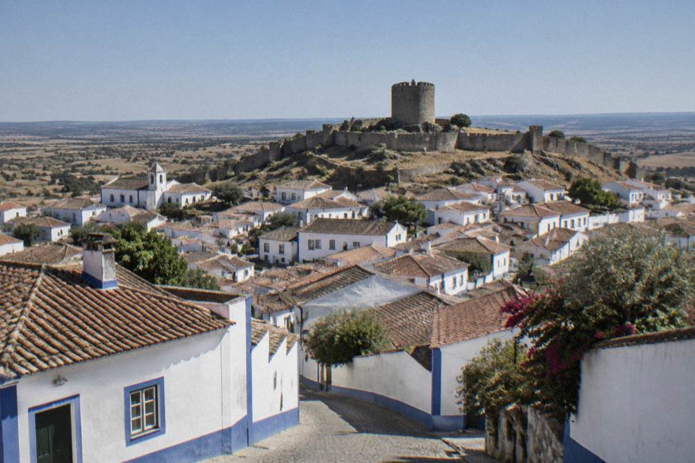 A round stone castle keep standing on a hilltop above low white town rooftops