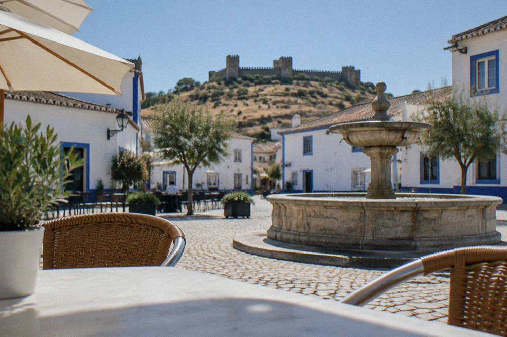 A quiet town square with white buildings, café tables, and a stone fountain in late afternoon light