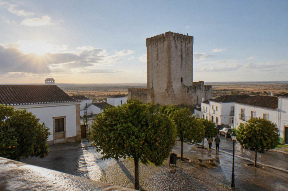 A stone keep tower seen from a cobbled square with orange trees and low white buildings