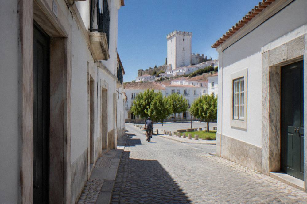 A narrow street in Estremoz with white marble doorframes and lintels set into whitewashed houses, a cyclist passing in the middle distance.