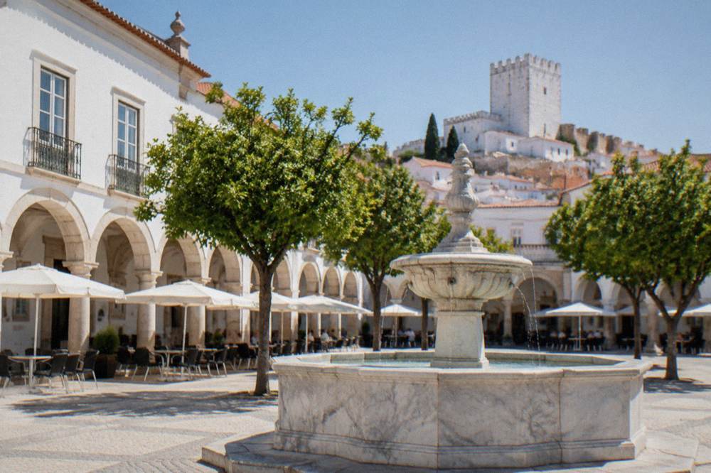 An arcaded square in Estremoz with a marble fountain at its centre and cafe tables under the shade of the arches.