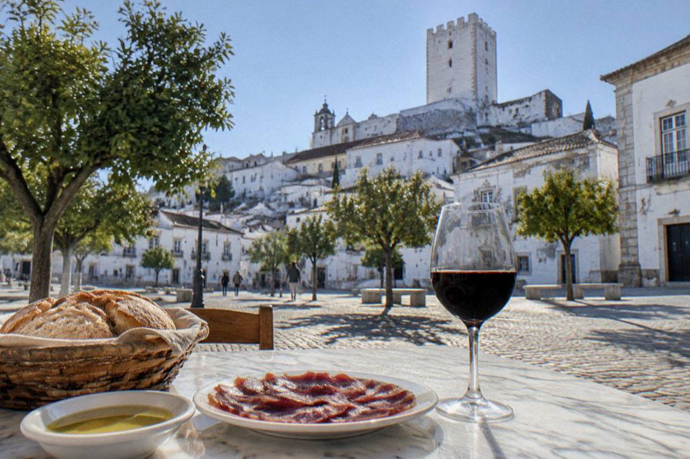 A rustic Alentejo table with bread, olive oil, a plate of cured pork and a glass of red wine on a white marble surface.