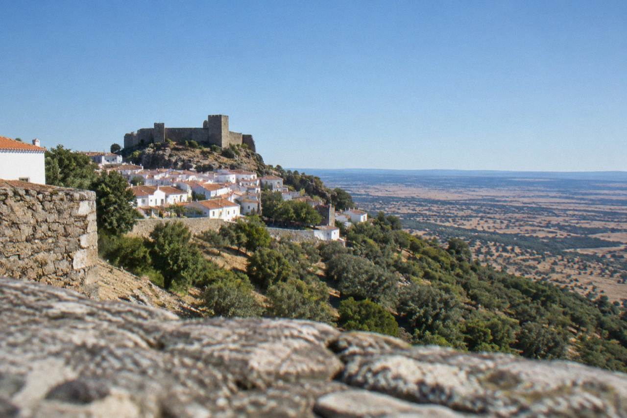 A walled hilltop village with a stone castle rising above wooded ridges under a wide sky