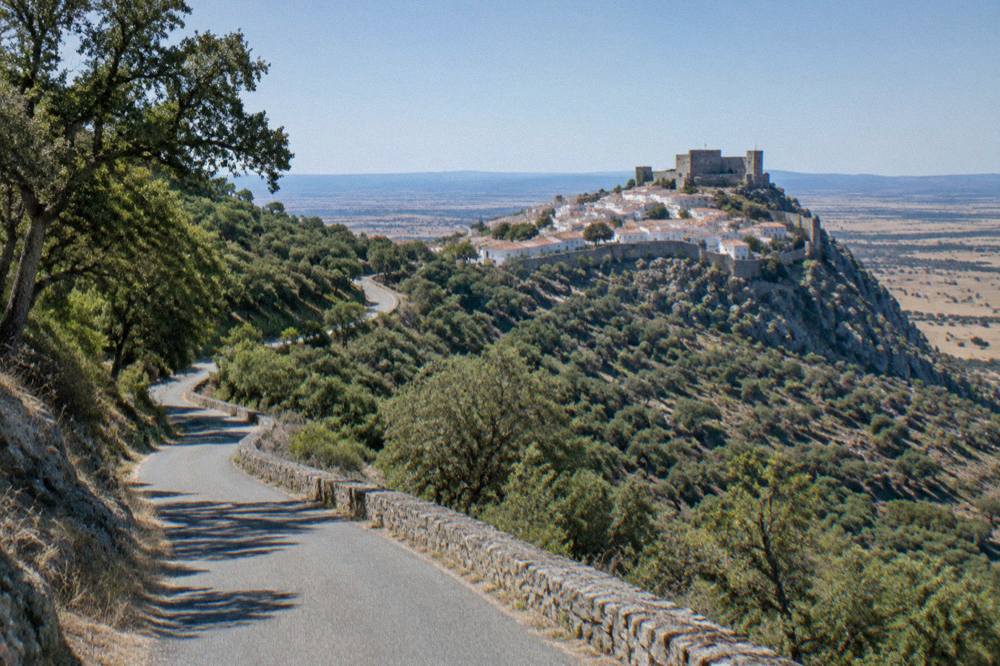 A country road climbing through wooded hills toward a distant walled village on a ridge