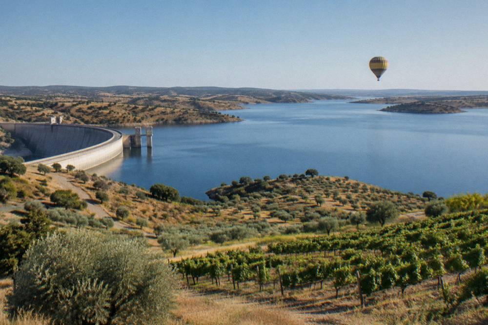 A hot air balloon drifting low over vineyards and the Alqueva reservoir at dawn