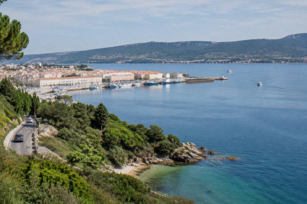 A coastal road winding through green hills above a small cove with clear water below