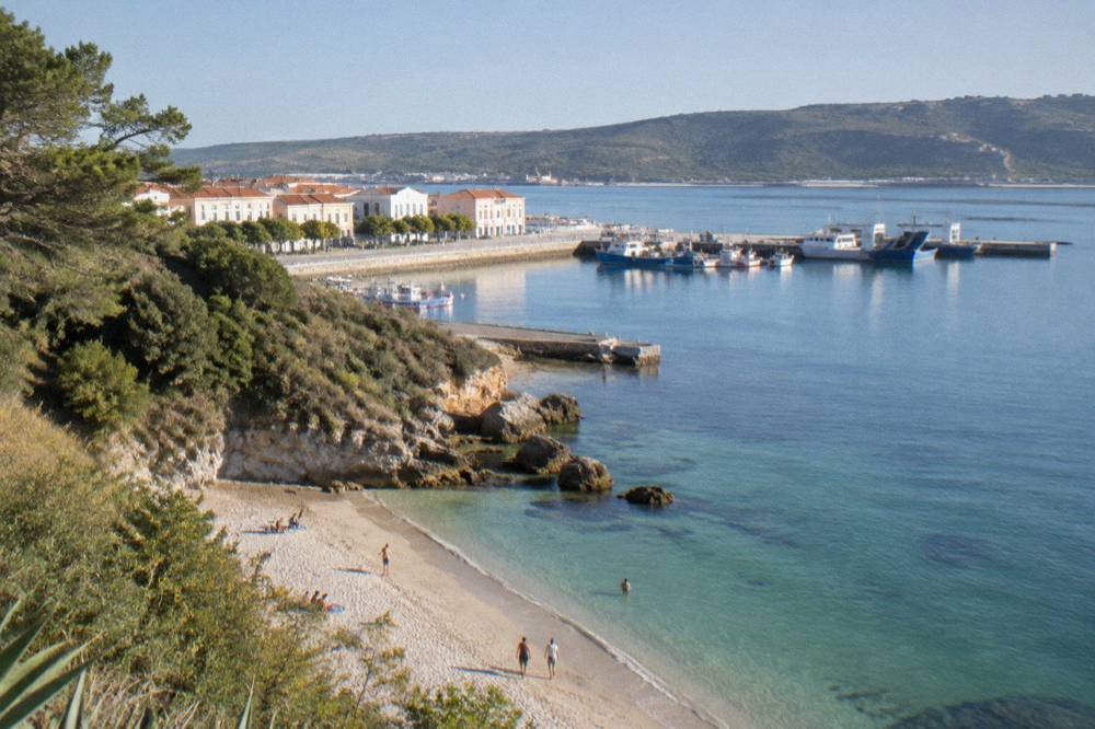 A small stone cove beach beneath wooded cliffs, with calm turquoise water and a handful of people on the sand