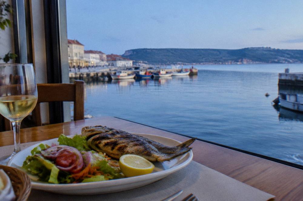 A grilled whole fish with lemon and salad on a restaurant table beside a window overlooking a harbour