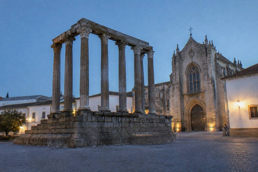 Roman temple columns standing in a square beside a Gothic church at dusk