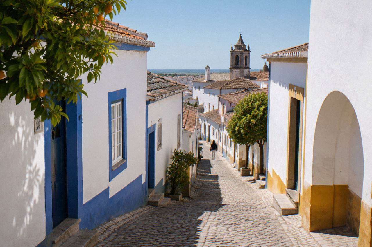 A narrow whitewashed street inside the walled historic centre of Évora with blue-painted door frames and a church tower rising over tiled rooftops