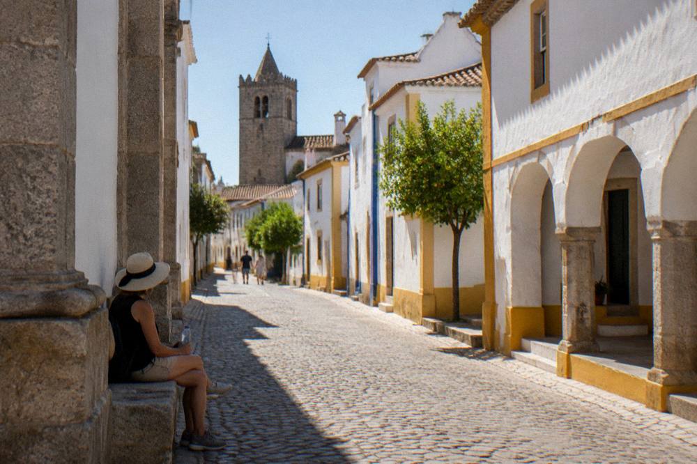 A visitor with a sun hat and water bottle resting in the shade of a church wall on a hot street in Évora