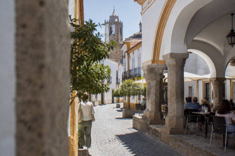 Shaded arcade along a square in Évora with tables and people resting out of the midday sun