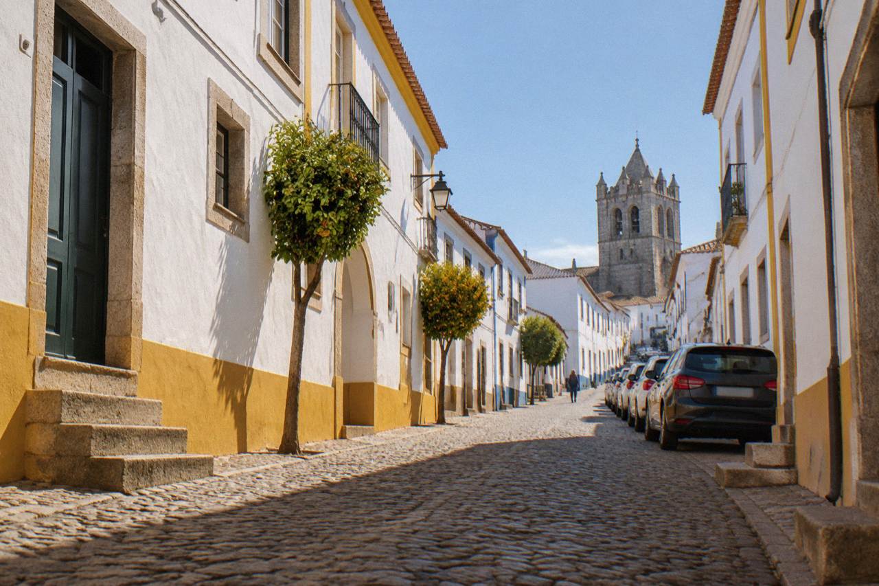 A narrow cobbled street inside the walled centre of Évora with parked cars lining one side and whitewashed houses above