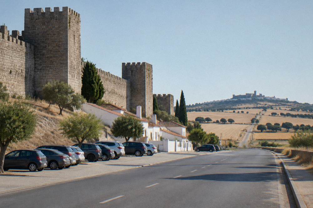 A country road crossing the Alentejo plain toward Monsaraz with wheat fields and a hilltop village on the horizon