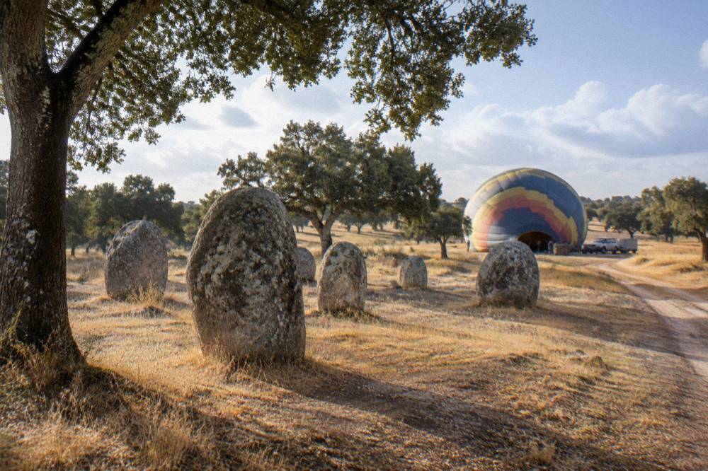 A balloon crew inflating an envelope on open ground beside cork oak trees in early morning light