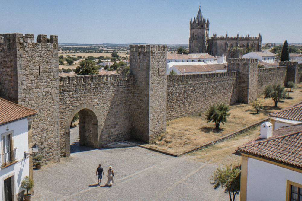 Two walkers on a cobbled square inside medieval city walls with a Gothic cathedral tower behind