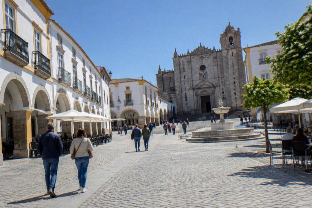 Visitors in light jackets walking across the Praça do Giraldo in Évora on a bright spring day