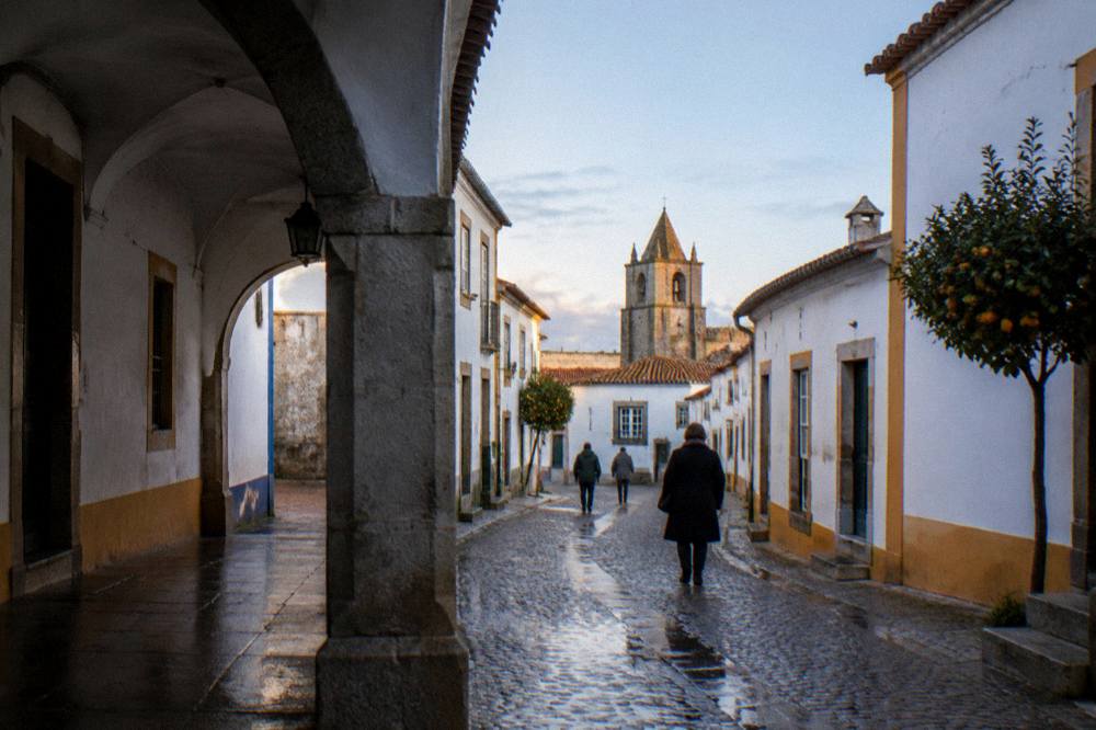The cloister arcade of a Évora monastery church in soft rain light, with wet stone paving reflecting the arches