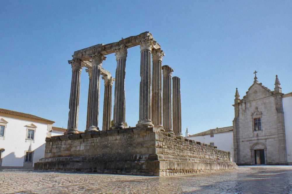 A sunlit square in Évora with the Templo Romano columns rising against a clear blue sky