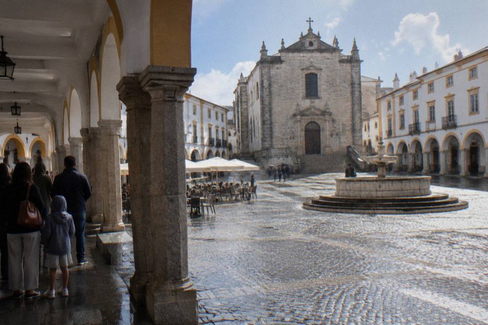 Families and visitors sheltering under an arcade on the Praça do Giraldo in Évora during light rain