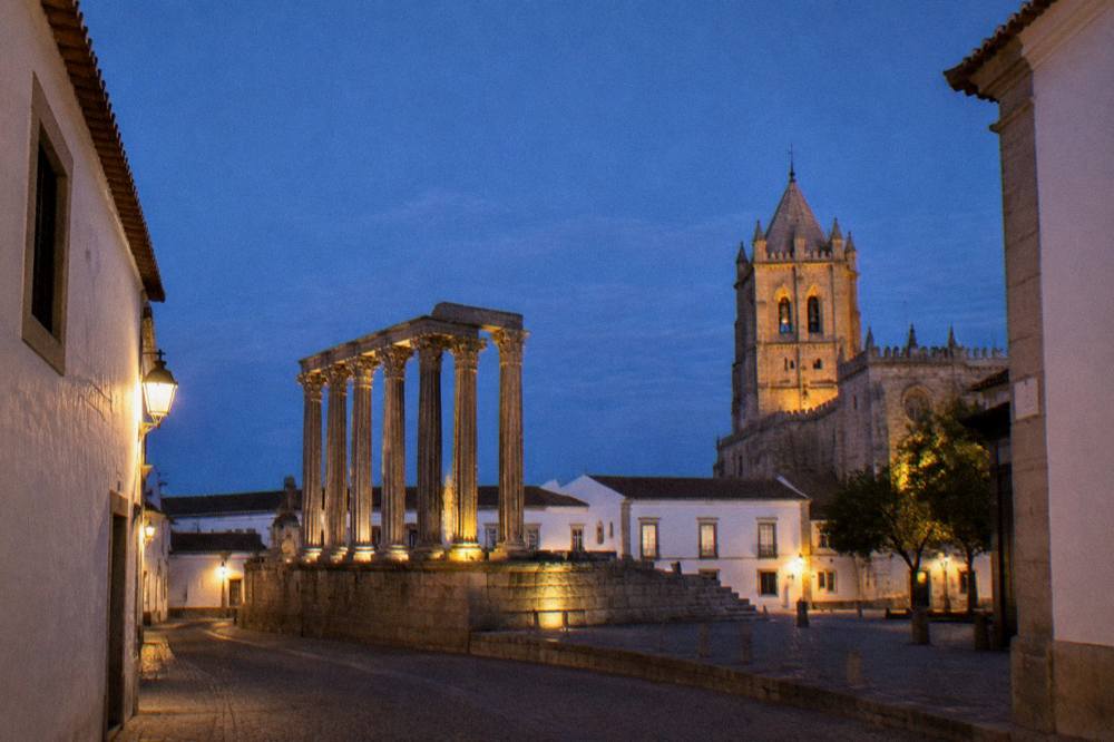The floodlit Roman temple and cathedral tower of Évora seen from a narrow street at night