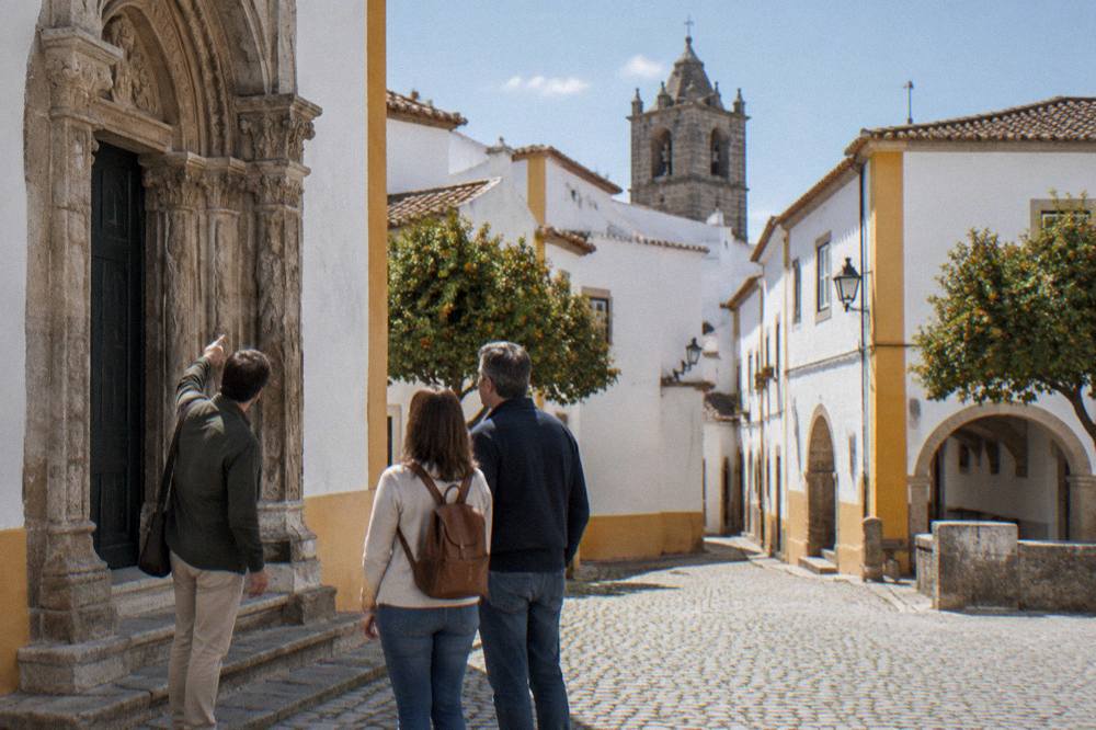 Guide pointing at a carved stone doorway while two visitors listen in a square in Évora