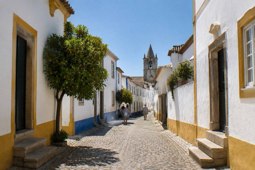 A sunlit cobbled street in Évora with white walls and blue and yellow trim under a deep blue summer sky
