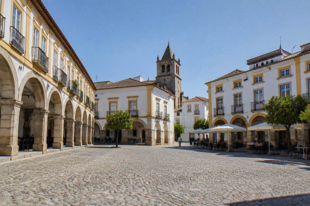 A wide cobbled square in Évora lined with arcaded golden stone buildings and café tables