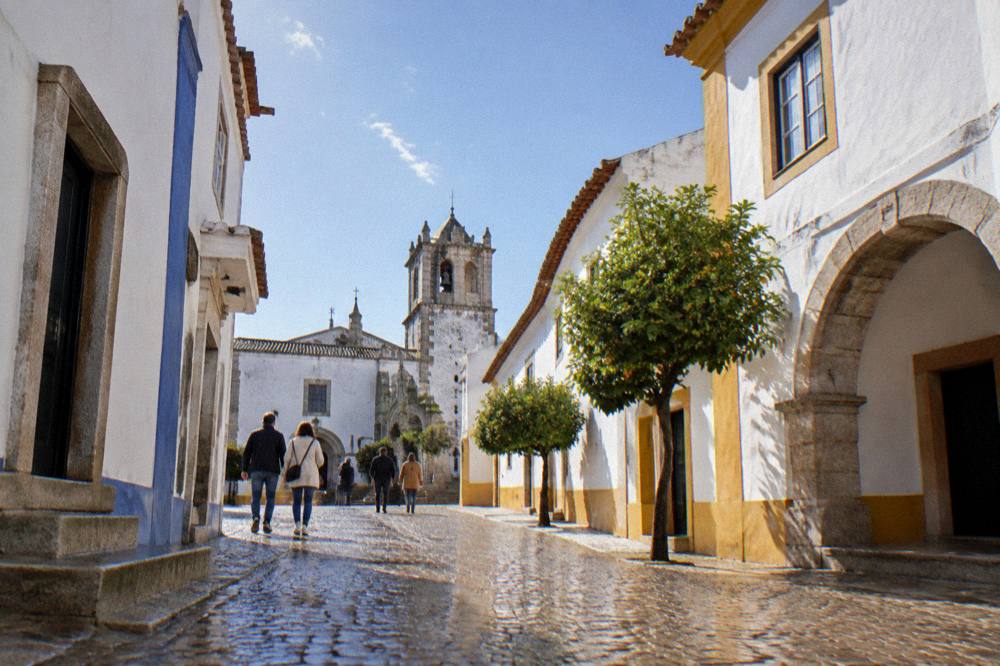 Visitors walking across a sunlit square toward the arches of a church in Évora