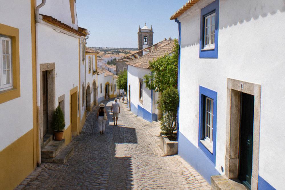 A shaded cobbled street with white walls and blue trim in Évora under a summer sky