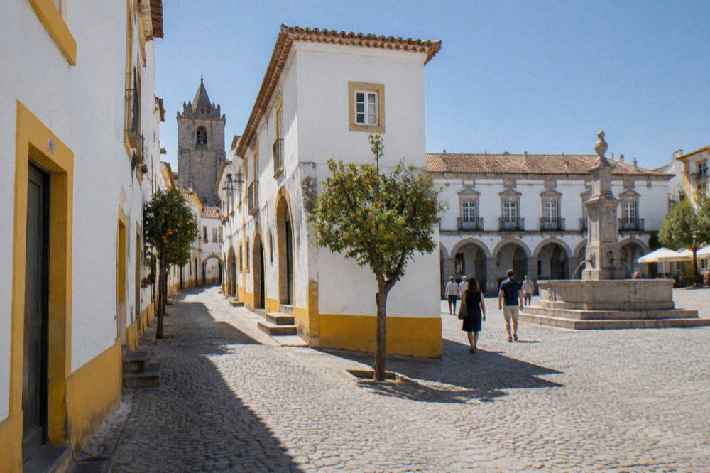 Visitors walking across a sunlit square in Évora beside old stone buildings with a fountain