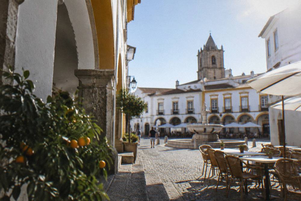 The arcaded main square of Évora with café tables, whitewashed buildings and the fountain in daylight