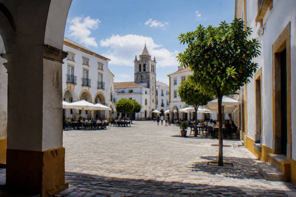 A wide granite-paved square lined with white arcade buildings and outdoor café tables