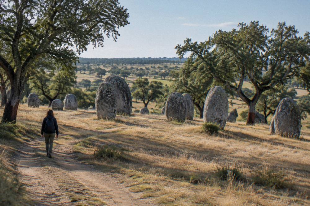 A visitor walking a dirt track toward standing stones on a wide treeless hillside in the late afternoon