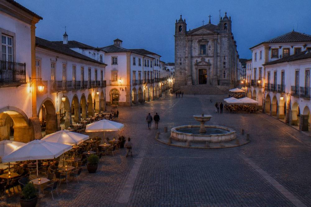 Arcaded square of Praça do Giraldo in Évora at dusk with café tables and warm lamplight