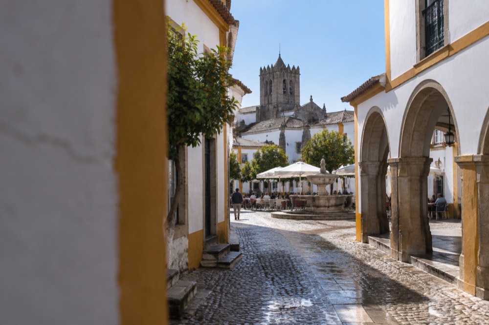 A sunny square in Évora with café tables under white arcades and a fountain